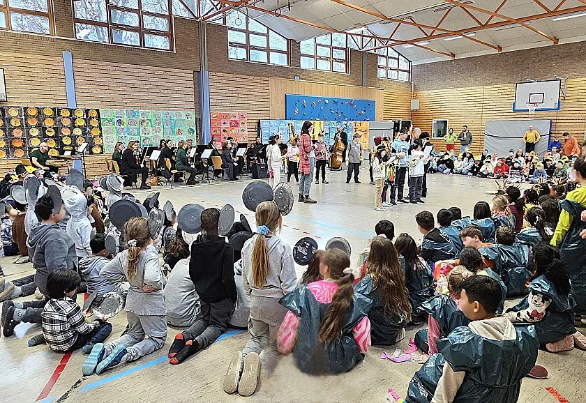 Schulkinder sitzen mit dem Ensemble der Bremer Philharmoniker im Kreis in der Sporthalle und verfolgen eine Aufführung einer Kindergruppe, die in der Kreismitte steht.