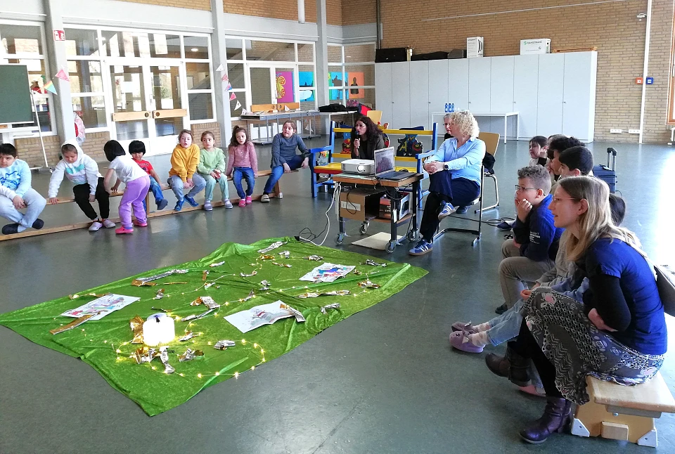Kinder lesen 3 Frauen und Mitsch&uuml;ler:innen im Halbkreis sitzend ein M&auml;rchen vor.