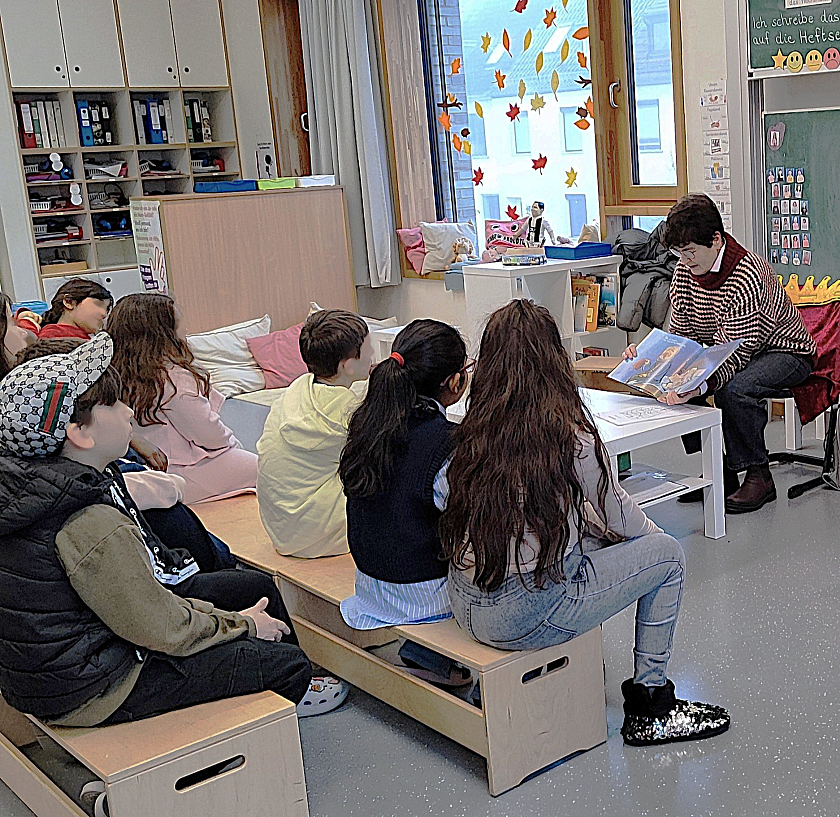 Kinder sitzen im Halbkreis und betrachten eine Bilderbuchseite, die ein Mitglied des Rotary Clubs in den Händen hält und den Kindern zeigt.