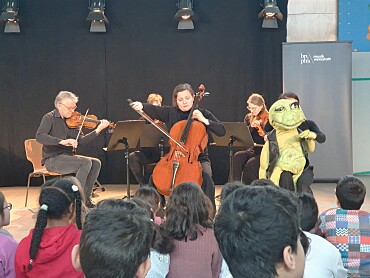 Schulkinder sitzen vor einem musizierenden Streichquartett der Bremer Philharmoniker und Eduardo, Puppe des Bremer Puppentheaters Mensch, Puppe.