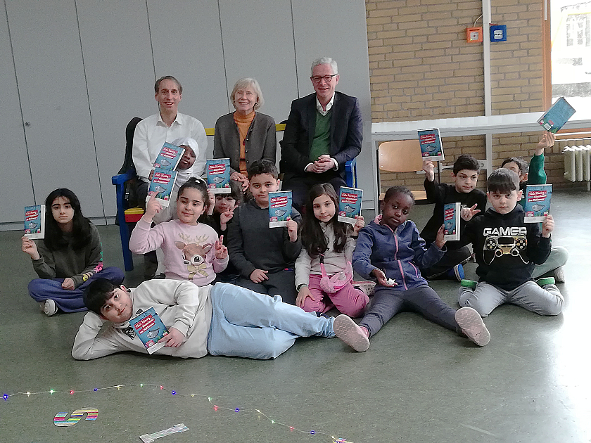10 Schulkinder sitzen auf dem Fußboden vor 3 Erwachsenen und halten ein Buchgeschenk in der Hand.