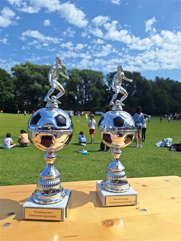 2 Wanderpokale der Fußballmeisterschaft von der Grundschule Pastorenweg stehen auf                     einer Holztischplatte. Im Hintergrund sind Kinder auf einem Fußballplatz zu sehen.